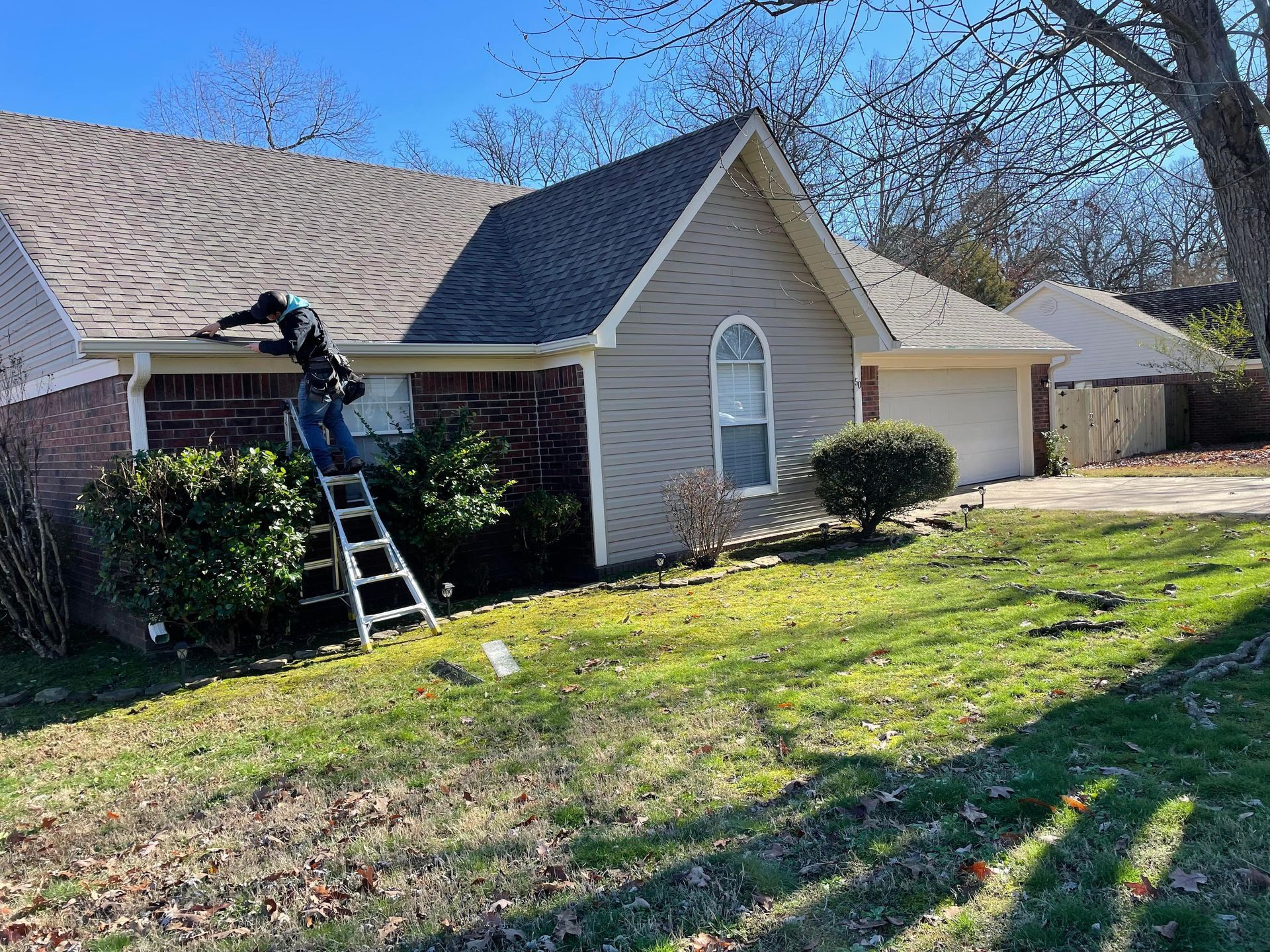 Worker on ladder repairing roof of beige house with gray shingles in a front yard.