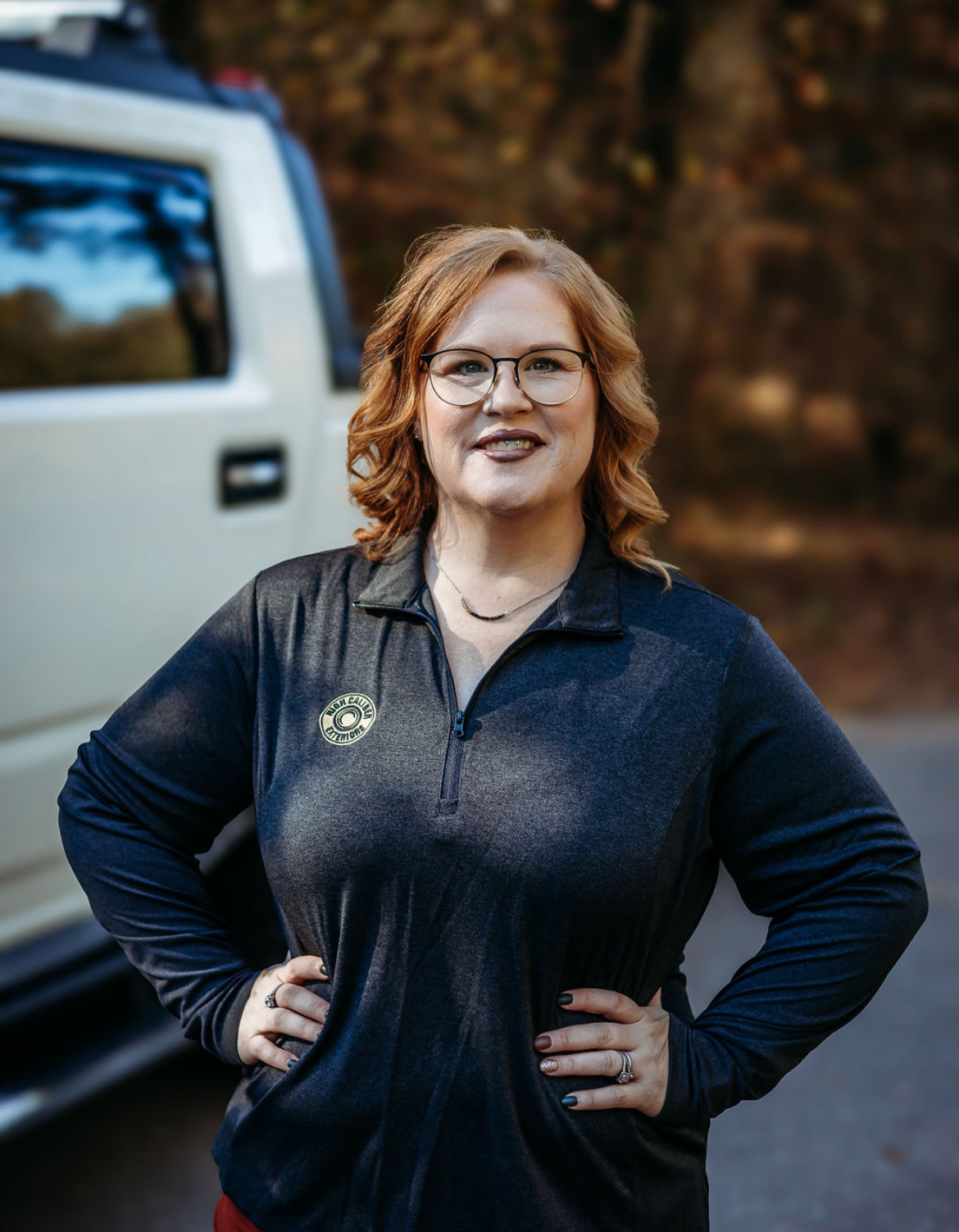 Smiling person with glasses in a dark jacket, standing hands-on-hips beside a white truck outdoors.
