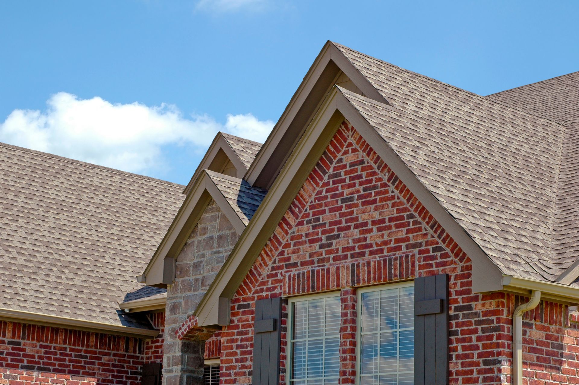 Brick house roof and gables with brown shingles under a partly cloudy blue sky
