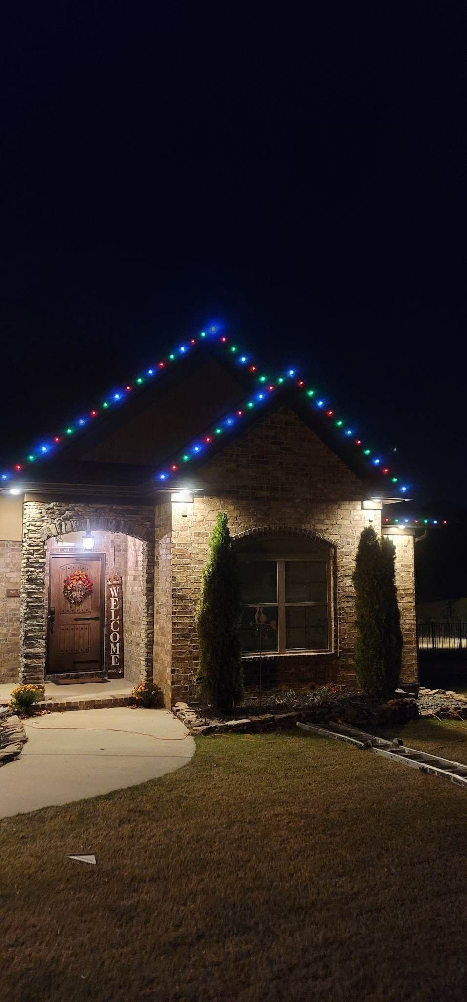 Small brick house at night with blue and white holiday lights along the roofline and a lit front porch