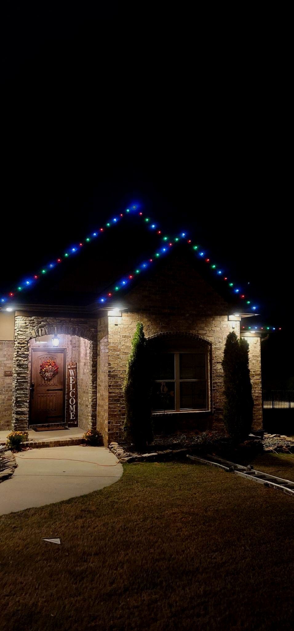 Brick house at night with blue roof lights and a lit front door, seen from the driveway.