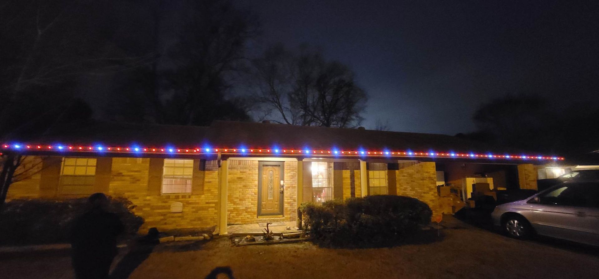 House with colorful holiday lights along the roof at night, with a parked car beside it.