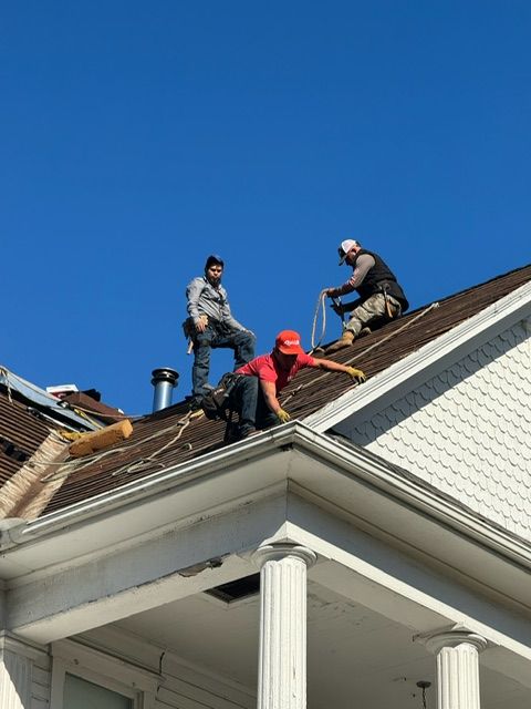 Three roofers working on a house roof under a clear blue sky, one in a red jacket.