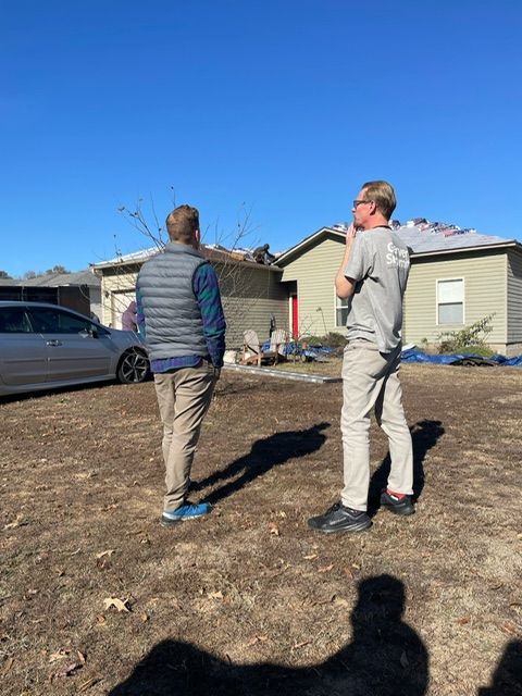 Two people standing in a bare yard near a house on a sunny day