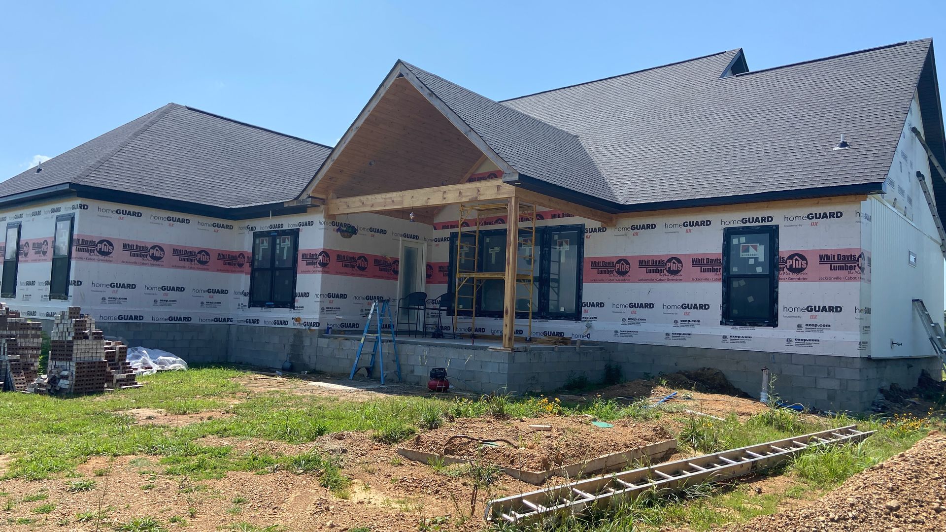 Partially built house with gray roof and exposed wrap, set on a grassy lot under bright sunlight