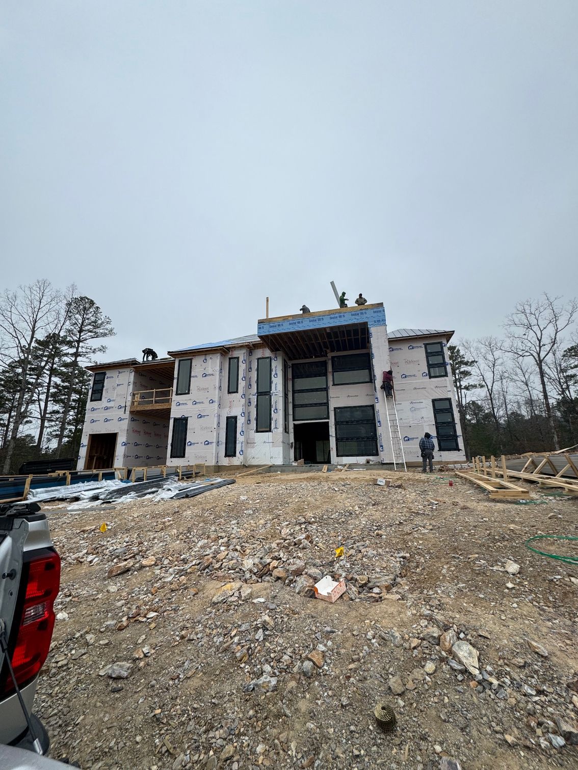 Unfinished multi-story house under construction on a gravel lot, with a parked red vehicle at left.