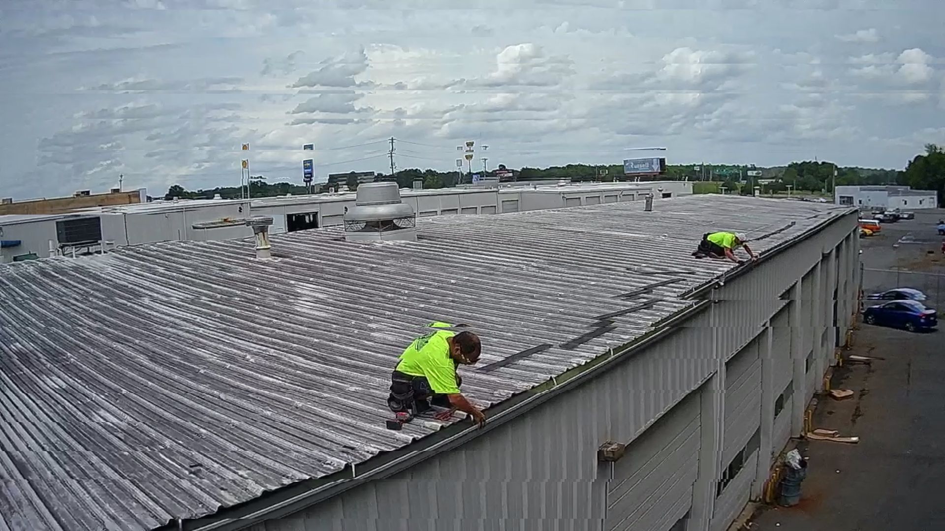 Workers in neon vests inspect a corrugated roof on a large industrial building under cloudy skies.