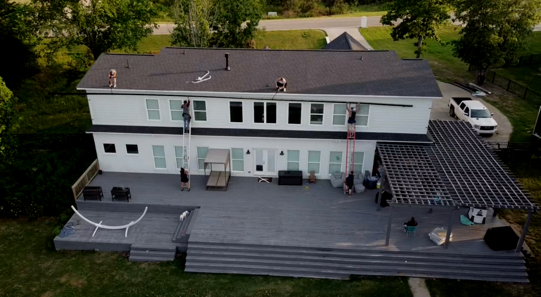 Aerial view of a white two-story building with a large wooden deck and outdoor seating, surrounded by trees.