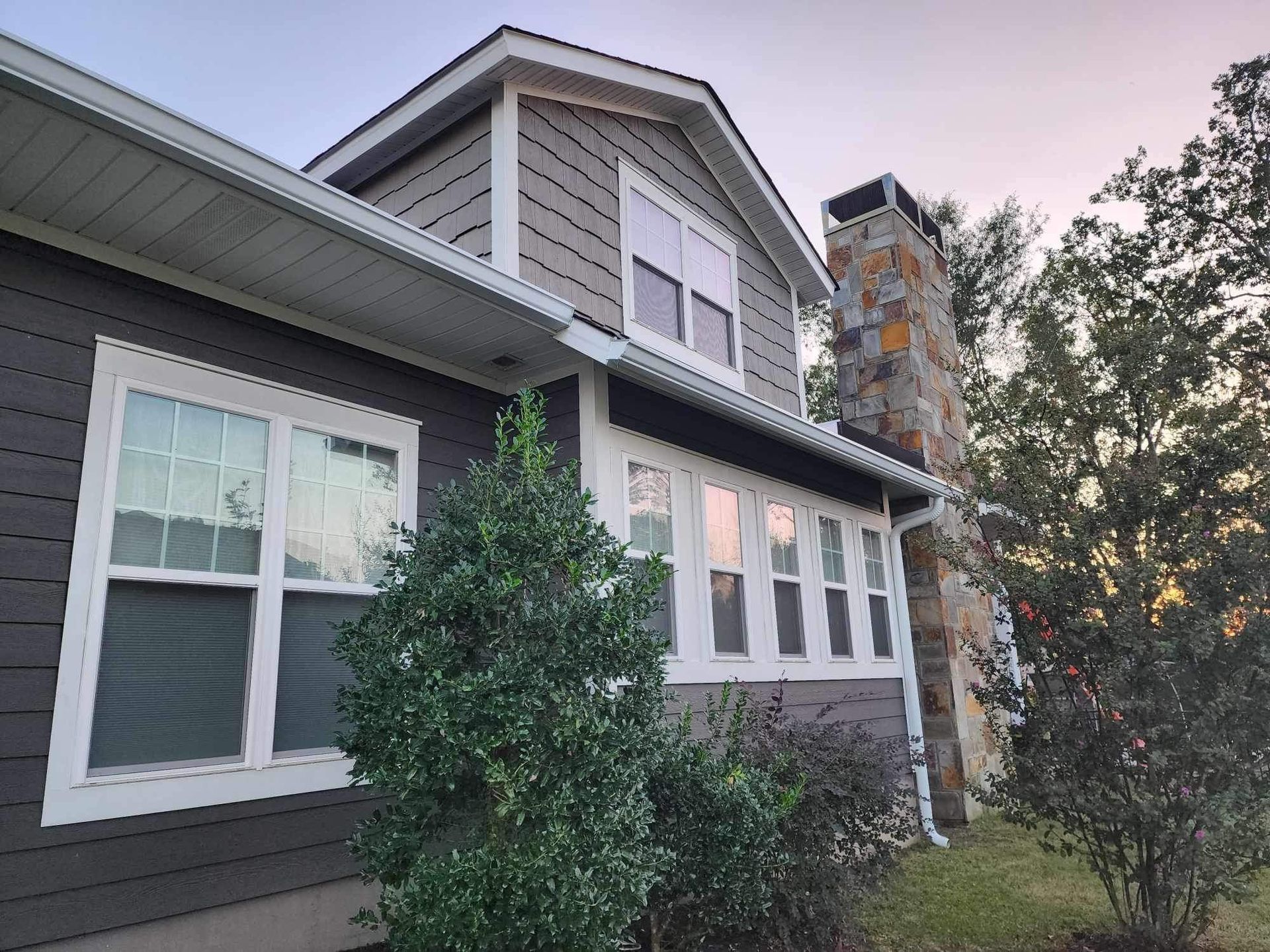 Gray house exterior with white trim, dormer windows, and a stone chimney at dusk