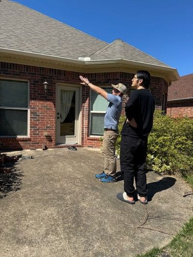 Two people standing outside a brick house, pointing toward the roof on a sunny day