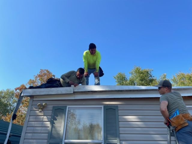 Three workers on a roof installing shingles above a house under a clear blue sky