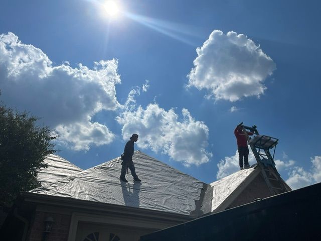 People on a roof under a bright sunny sky with clouds, one carrying equipment up a ladder