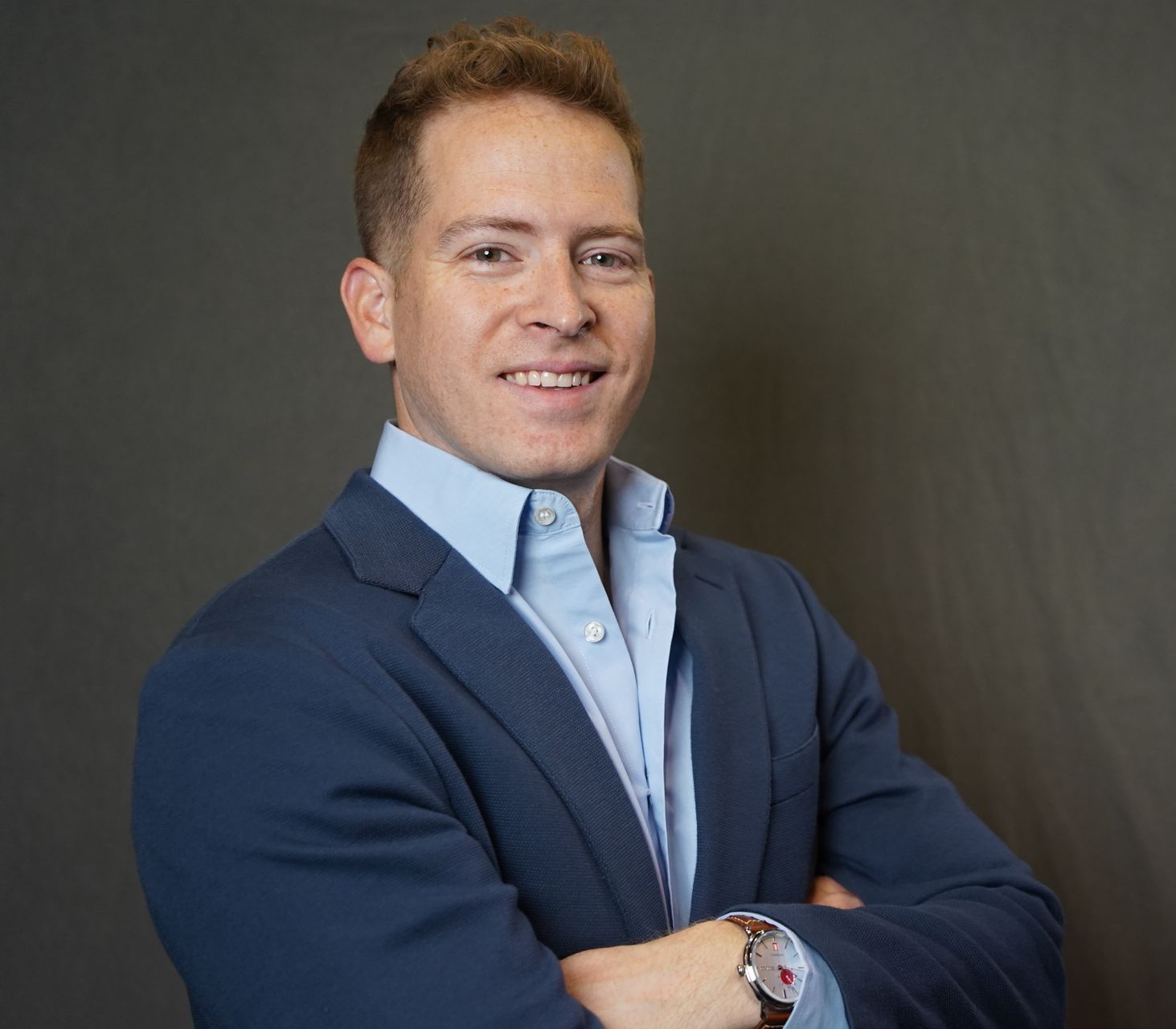 Smiling person in a blue blazer with arms crossed against a dark studio background