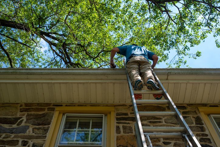 Worker on a ladder cleaning a roof gutter beside a stone house under green tree branches