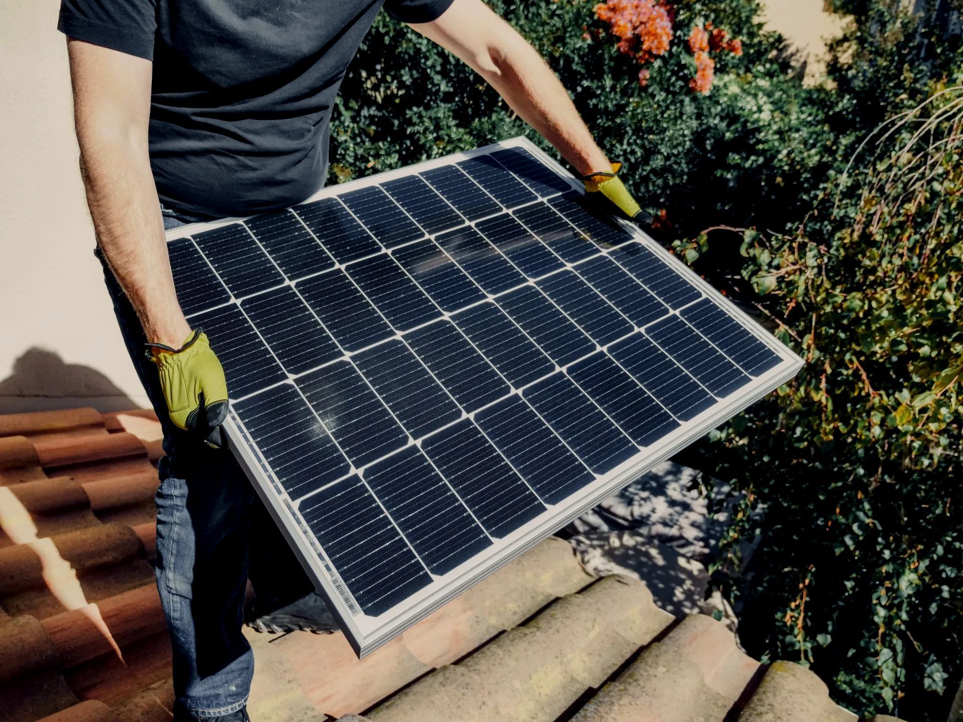 Person carrying a solar panel onto a tiled roof near bushes and flowers