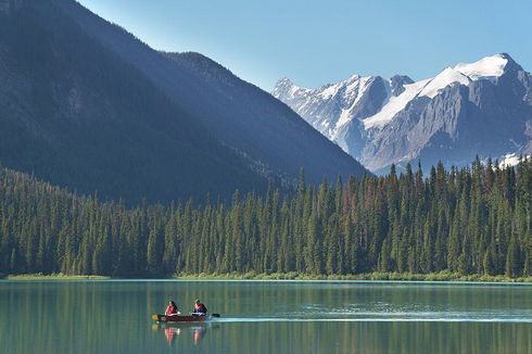 Two people canoeing on a calm mountain lake with pine forest and snowy peaks in the background