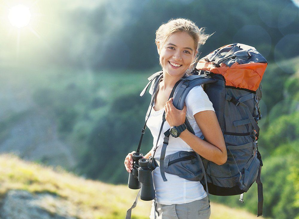 Eine Person mit Rucksack und Fernglas lächelt beim Wandern auf einem sonnenbeschienenen Bergpfad.
