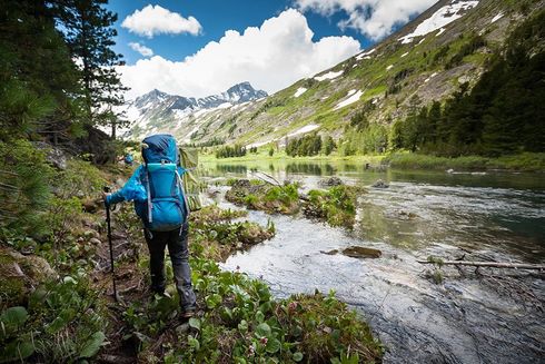 Hiker with blue backpack walking beside a mountain river in a green alpine valley.