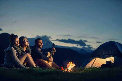 Friends sitting by a campfire near a lit tent at dusk in a mountain campsite