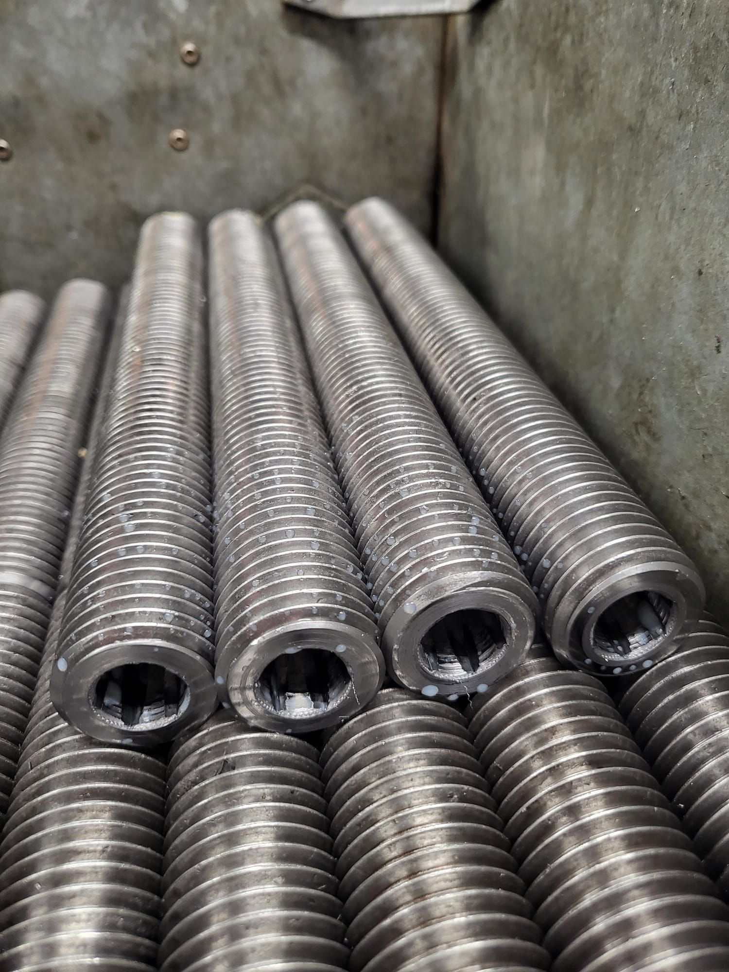 Close-up of a stack of cylindrical metal parts with threaded exteriors and hollow interiors, inside a metal container.
