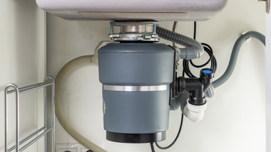 Garbage disposal unit under a kitchen sink, connected to plumbing and electrical wiring. Gray and white.
