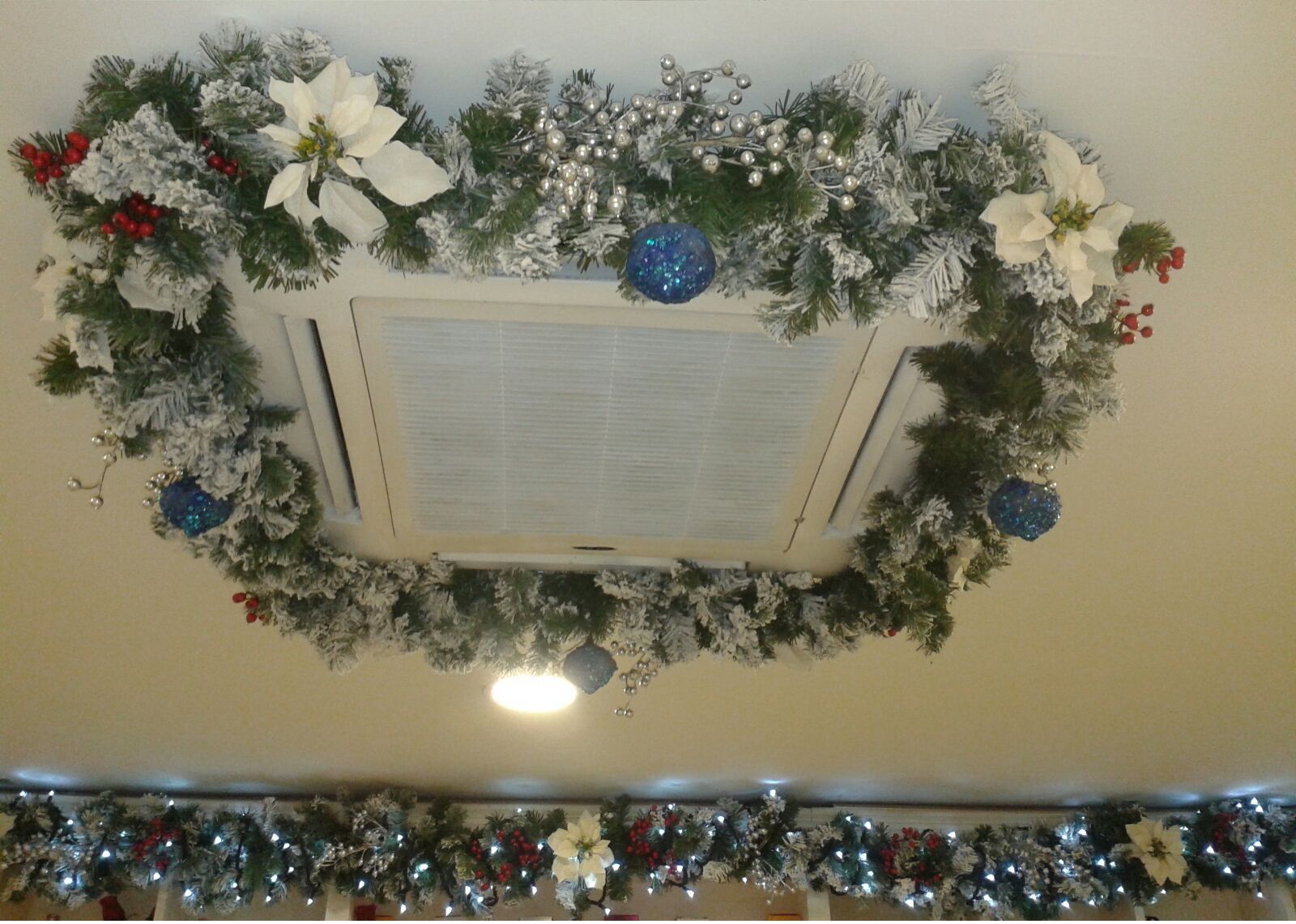 Ghirlanda natalizia avvolta attorno a una bocchetta di ventilazione sul soffitto, con fiori bianchi, ornamenti blu e bacche rosse.