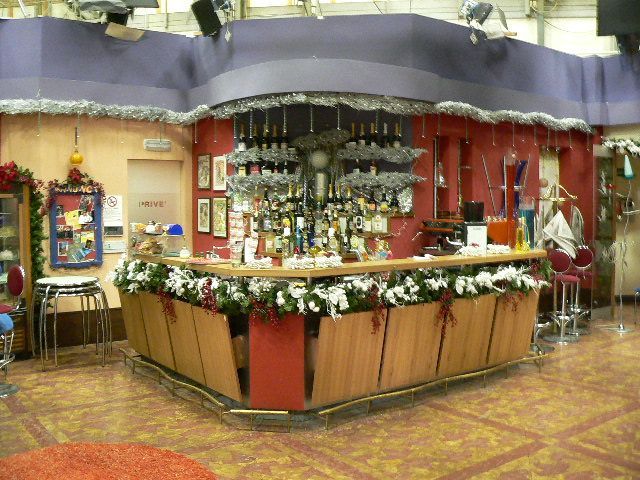 Bar al coperto con colorate decorazioni natalizie, bottiglie appese e un bancone in legno con ghirlande.