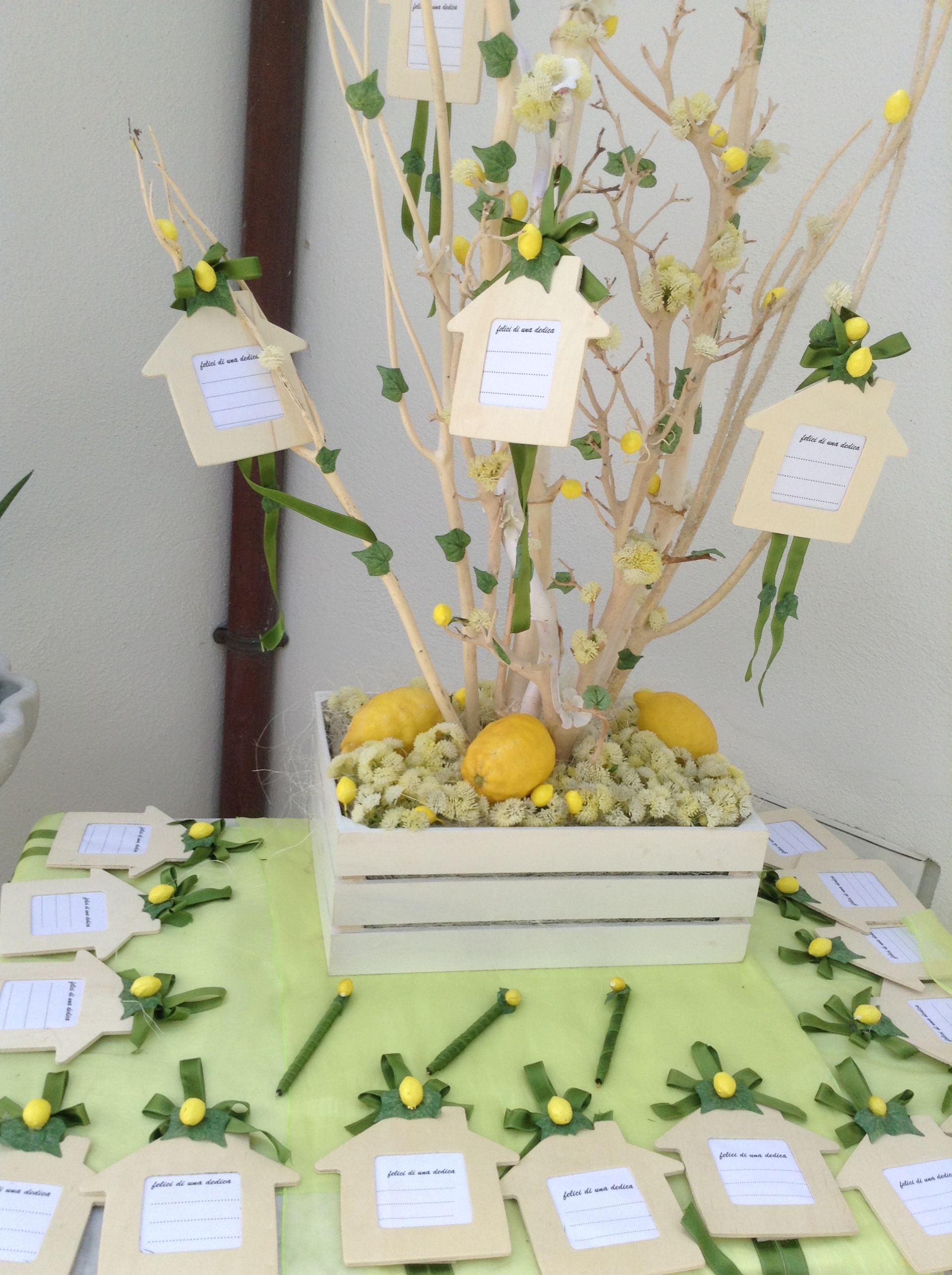 Composizione decorativa con fiori bianchi, dettagli color limone e cartoncini appesi su un tavolo ricoperto di verde.