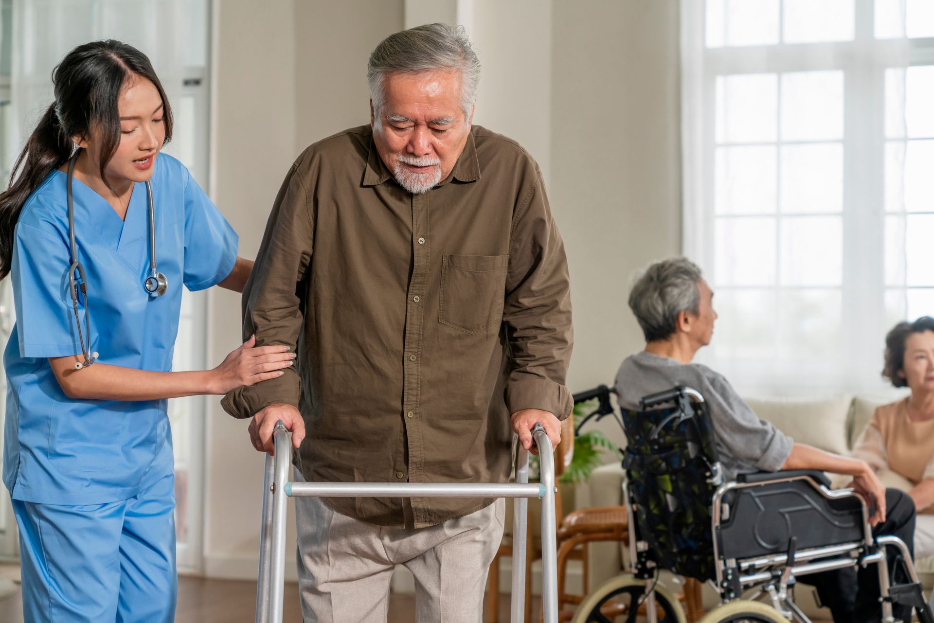 Caregiver helping an elderly man walk with a walker in a bright nursing home lounge.