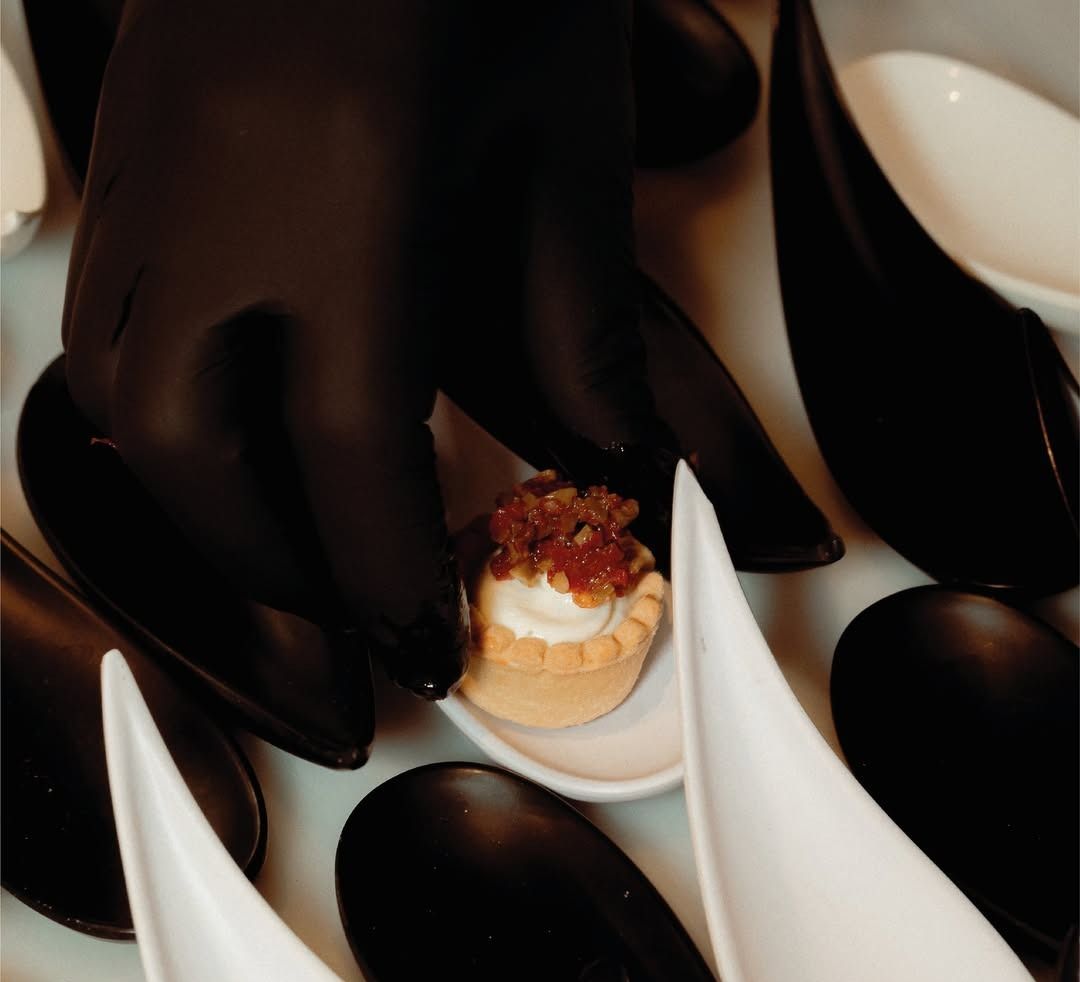 Una persona che indossa guanti neri tiene in mano un piccolo dessert su un cucchiaio