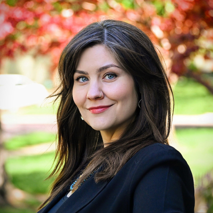 Smiling woman with dark hair in a black coat outdoors against a blurred red-and-green garden background