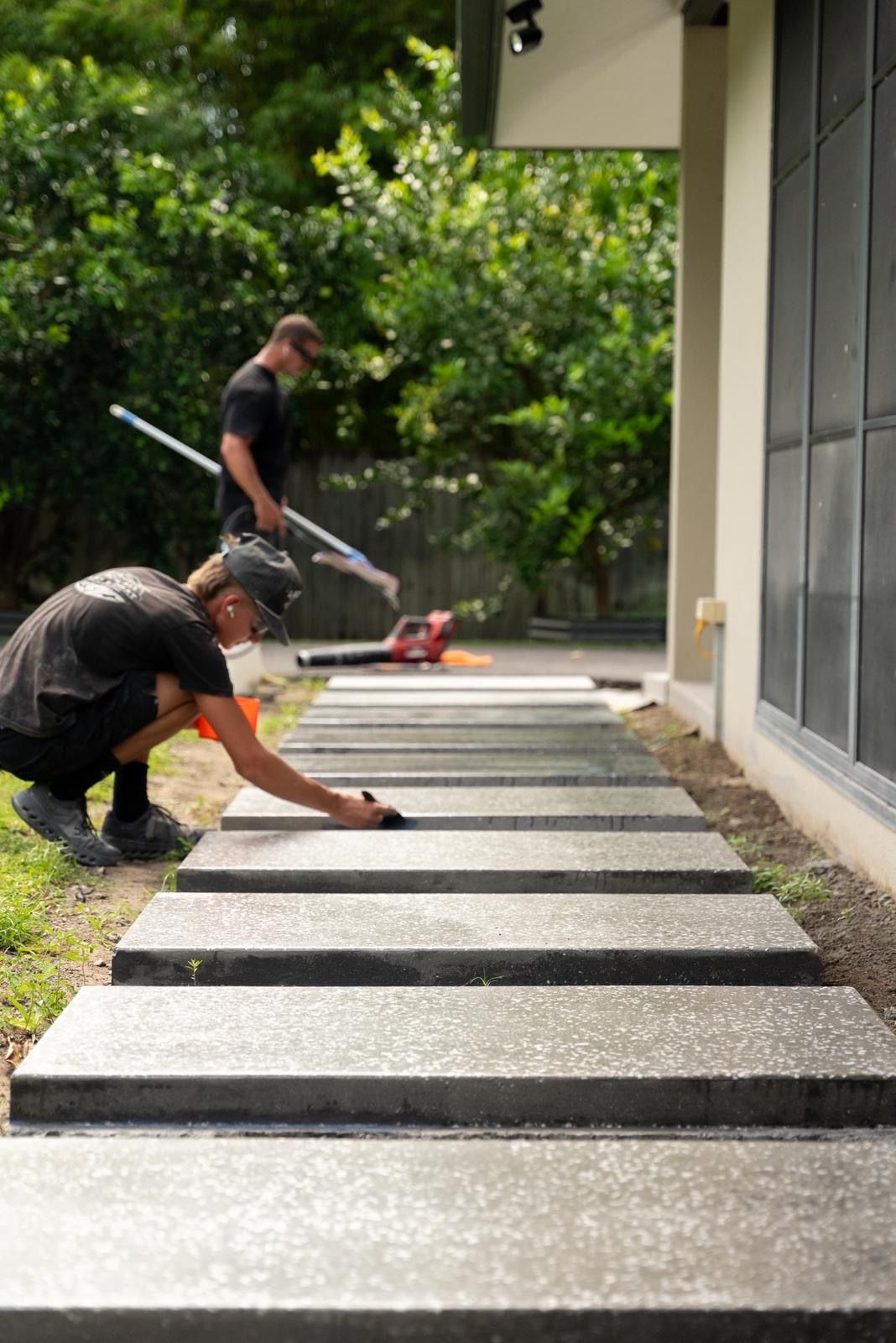 Two Workers Laying Gray Pavers Beside a Modern House in a Garden Path Area — All Grind & Coat in Cairns, QLD