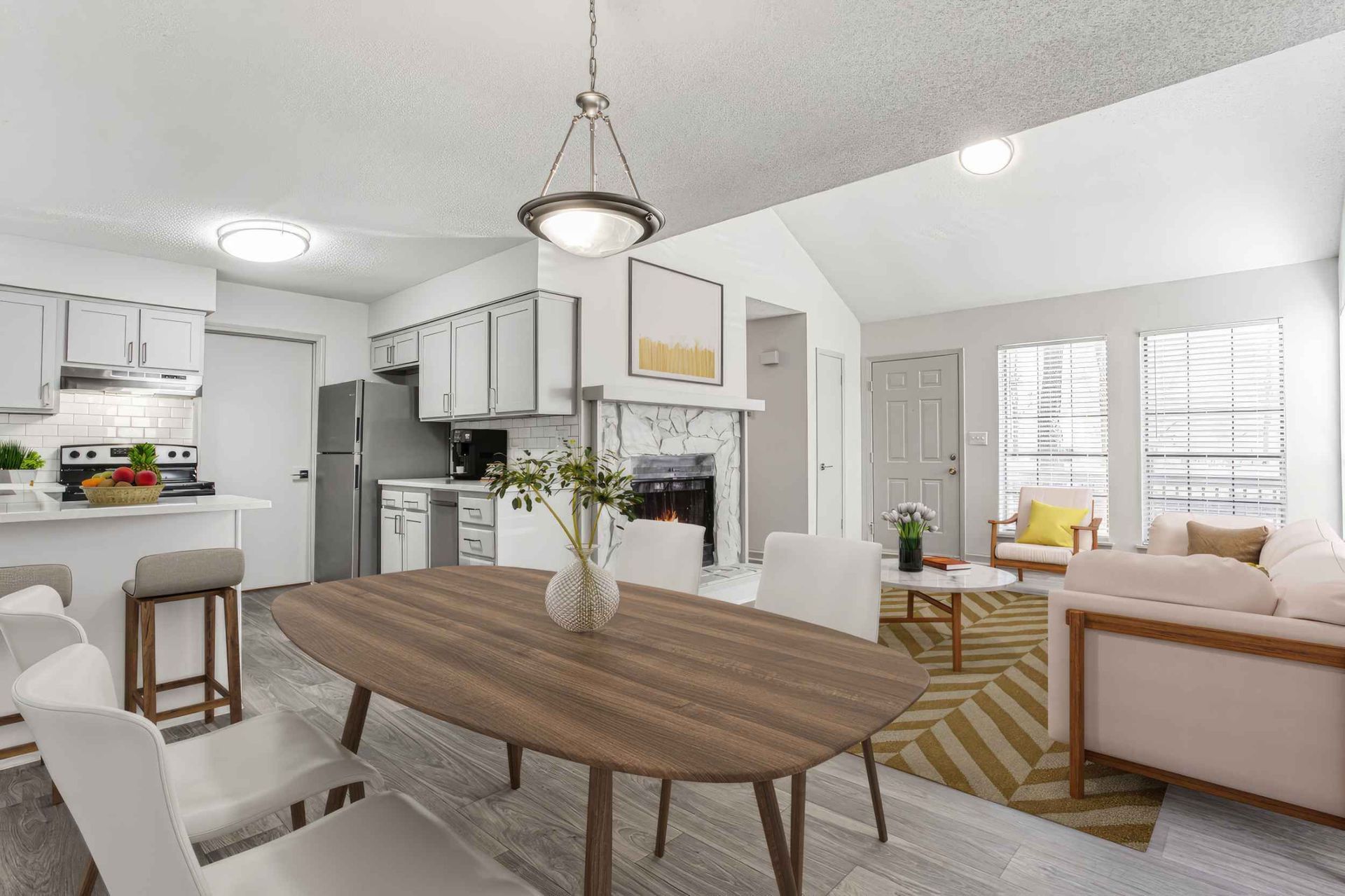 Bright open-plan living room and kitchen with wood dining table, white cabinets, and beige sofa by large windows