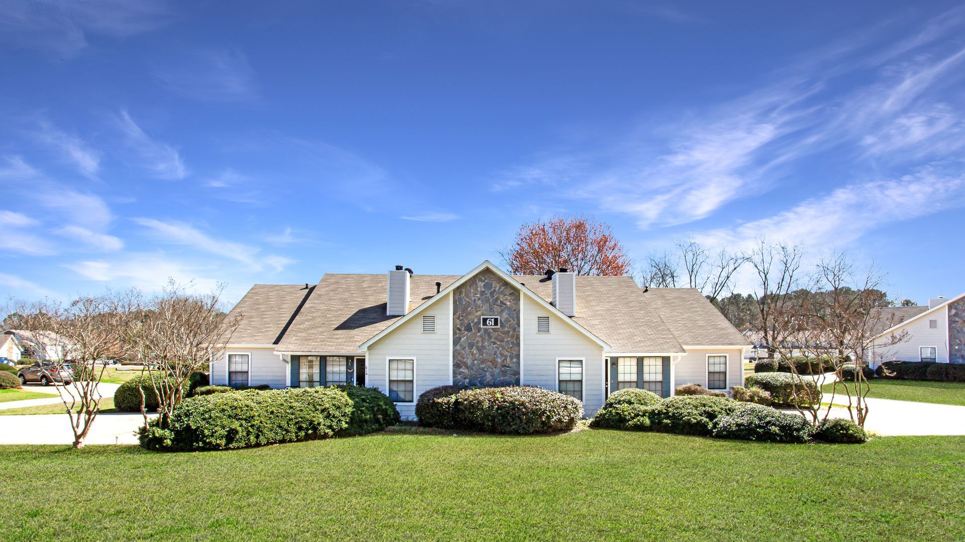 Suburban single-story house with a manicured lawn, shrubs, and a blue sky with clouds
