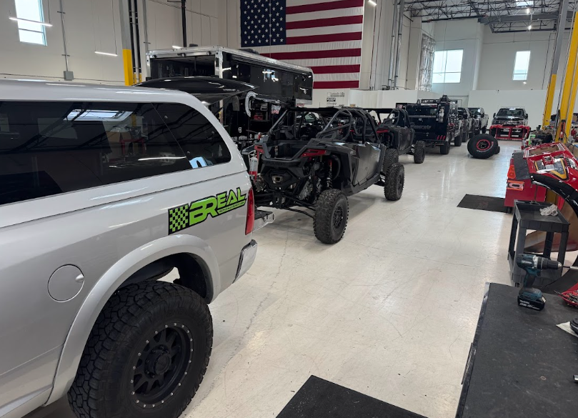 Off-road vehicles and trucks inside a garage, with an American flag on the wall.