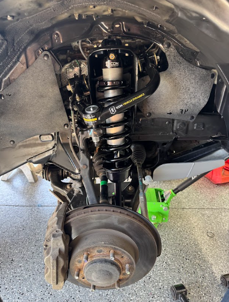 Car suspension and brake assembly with wheel removed, showing coil spring and exposed hub in a garage.
