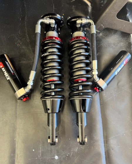Two black coilover shocks with red springs and two side-mounted gas shocks on a dark surface.