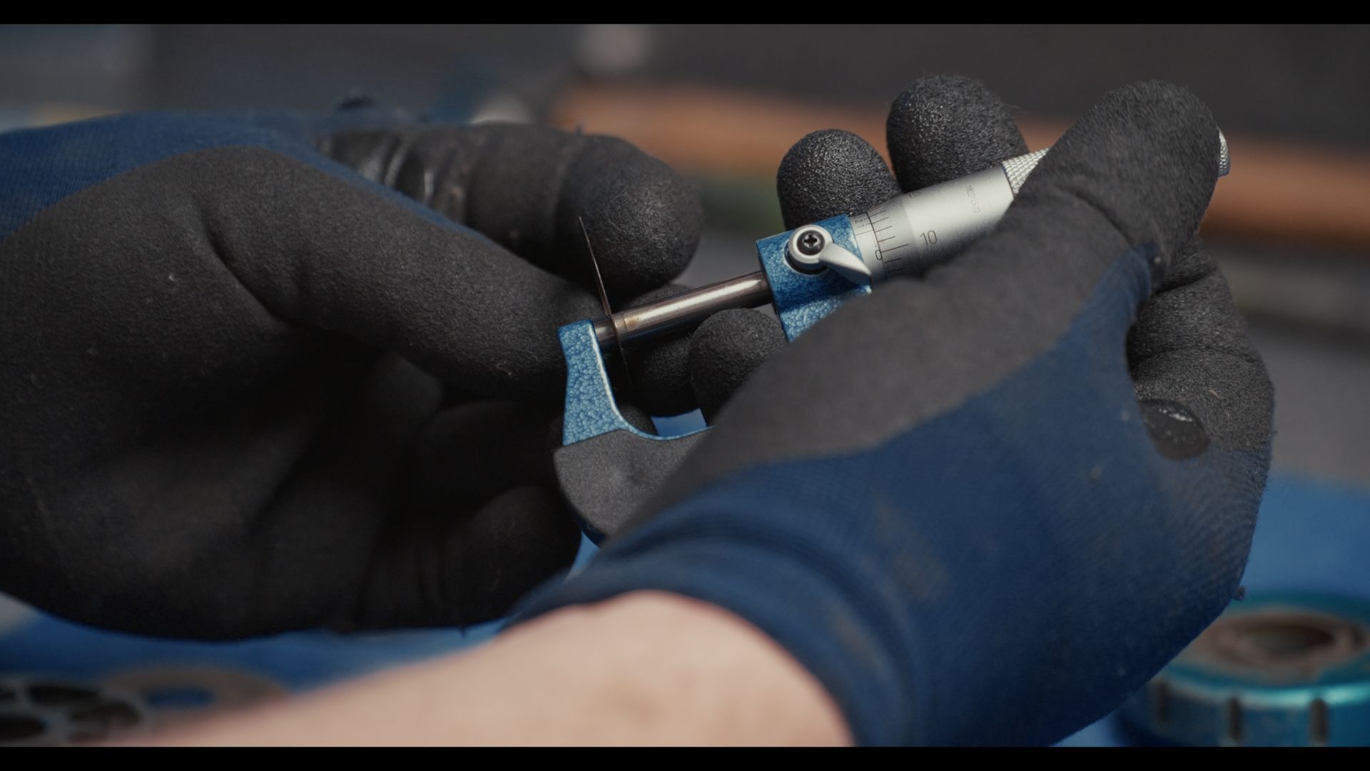 Gloved hands using a small wrench to adjust a metal part on a blue workbench