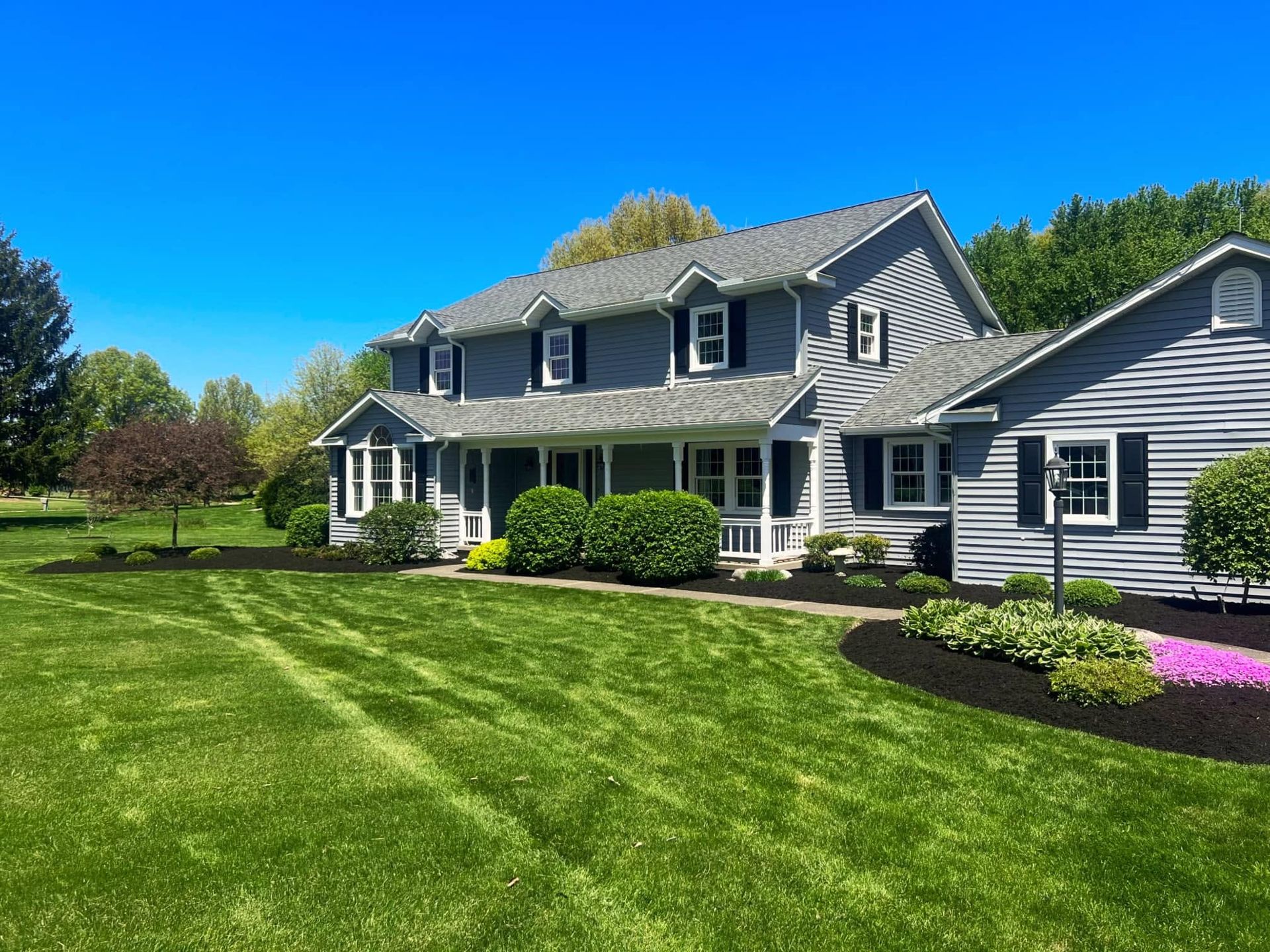 Large gray farmhouse with white trim and a manicured green lawn under a clear blue sky