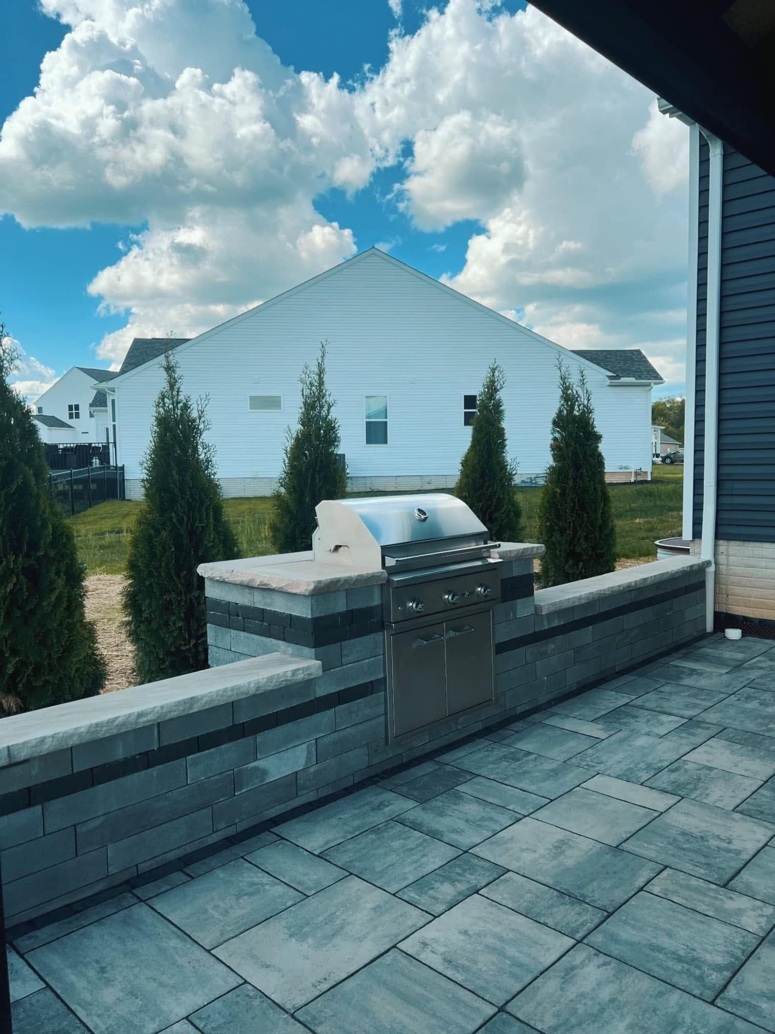 Patio with gray pavers, built-in grill, and landscaped yard beside a white house under a cloudy sky