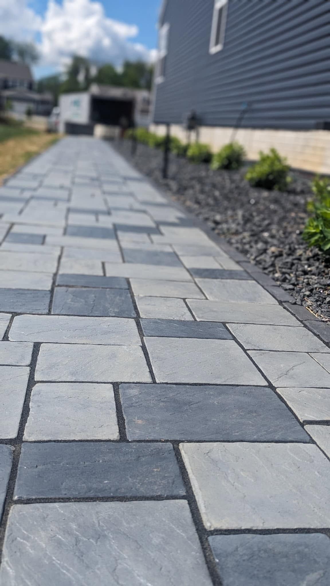 Stone paver walkway beside a gray house with black mulch and small shrubs