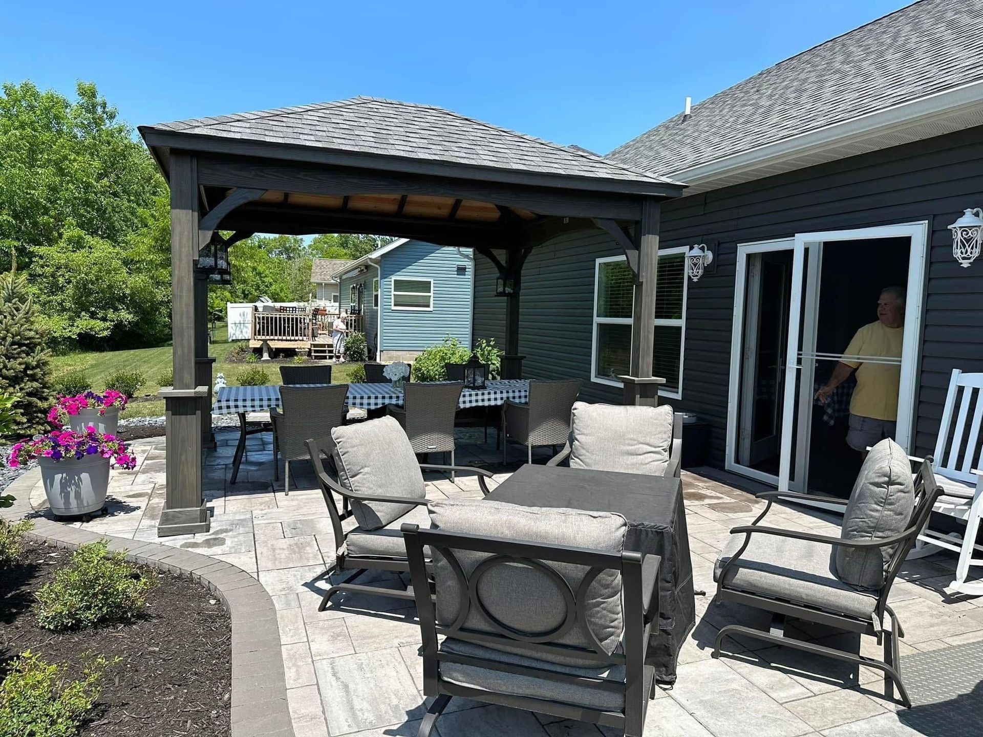 Patio with outdoor seating, stone pavers, and a covered grill beside a house in bright sunlight