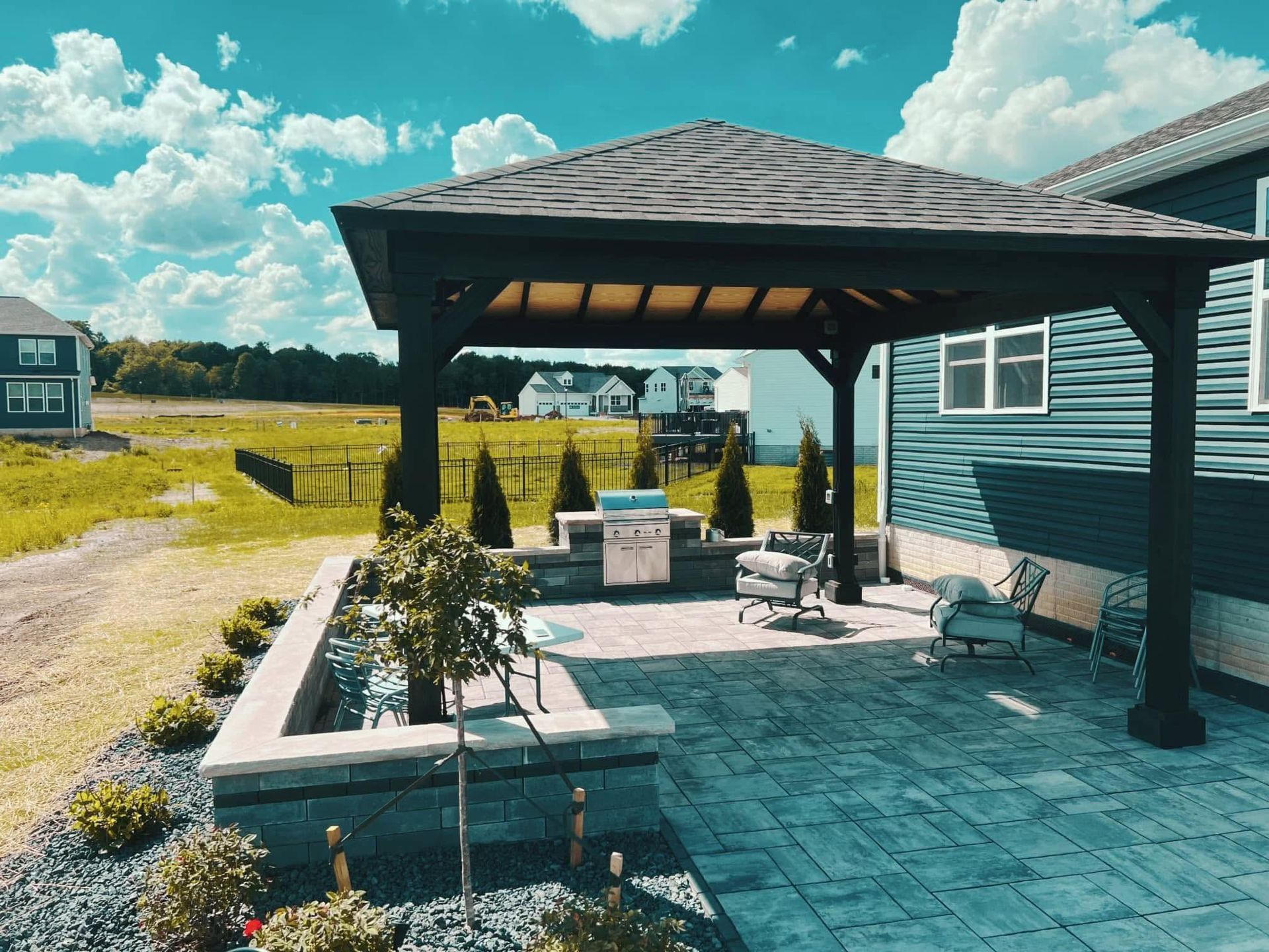 Covered patio with stone pavers, seating, and potted plants beside a house on a sunny day