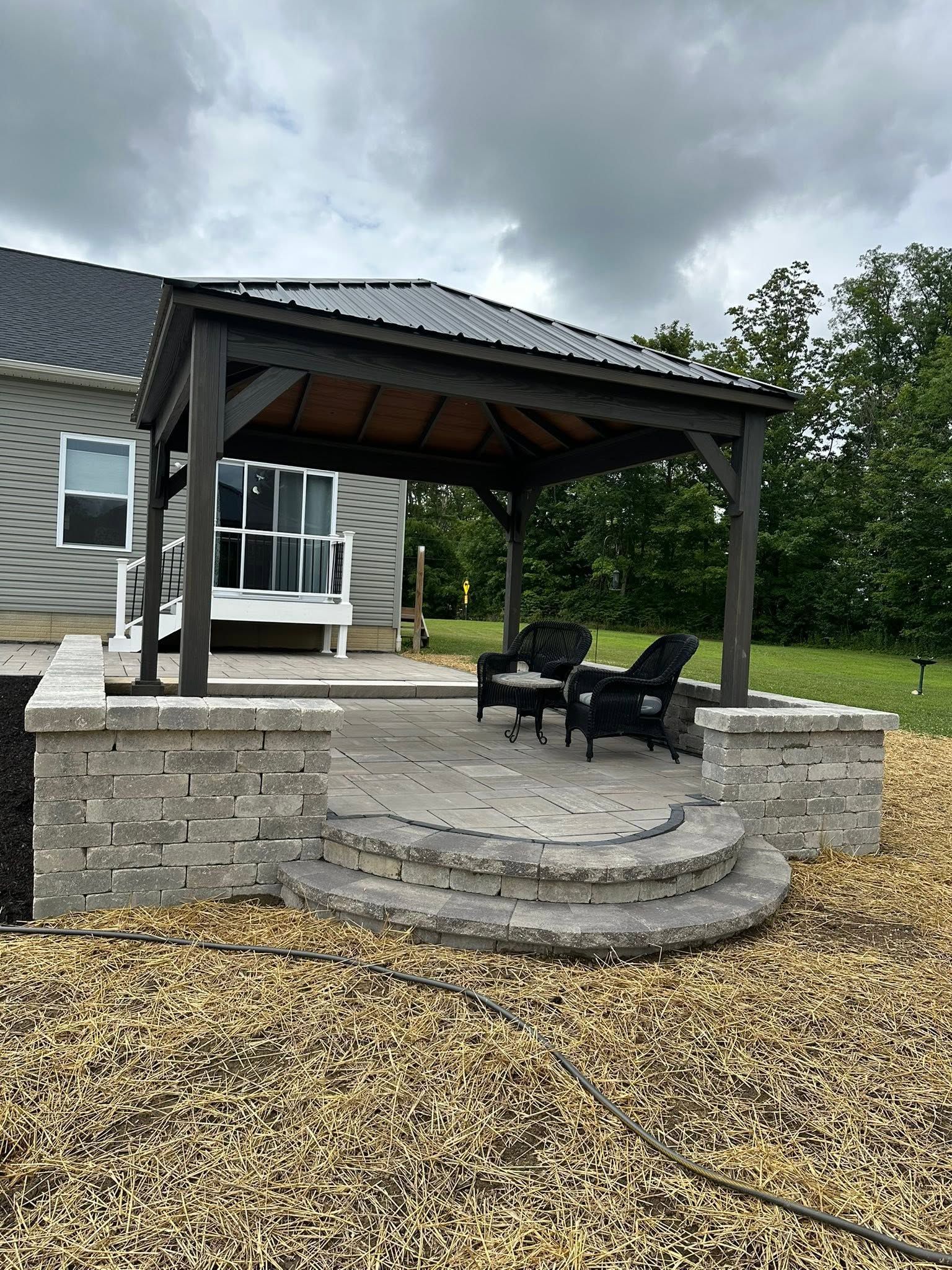 Stone patio with covered pergola, black chairs, and curved steps beside a house, under cloudy skies