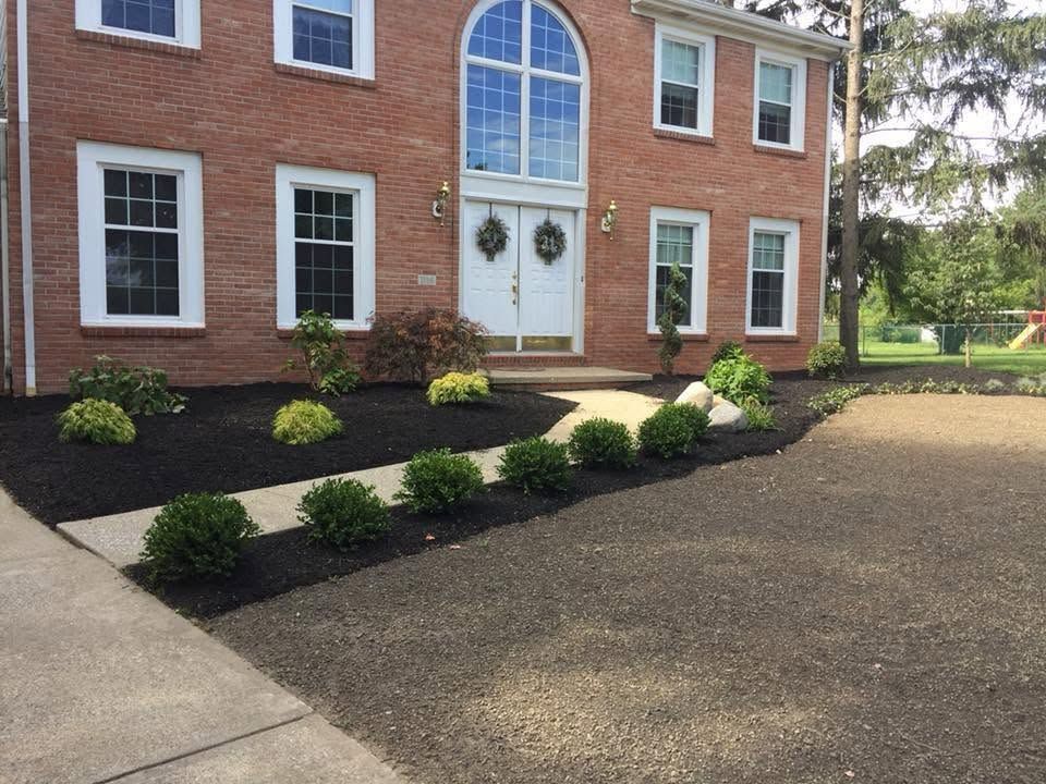 Brick house entrance with white double doors, arched window, and landscaped shrubs beside a driveway.