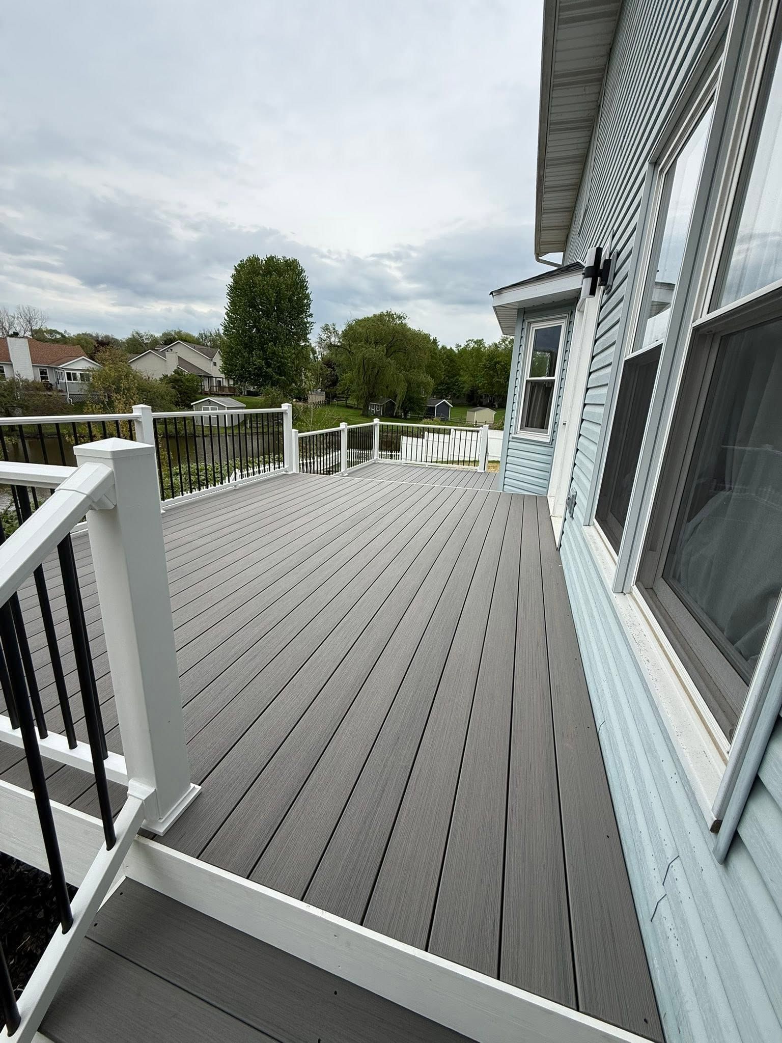Long gray deck beside a light blue house, with white railings and cloudy sky.
