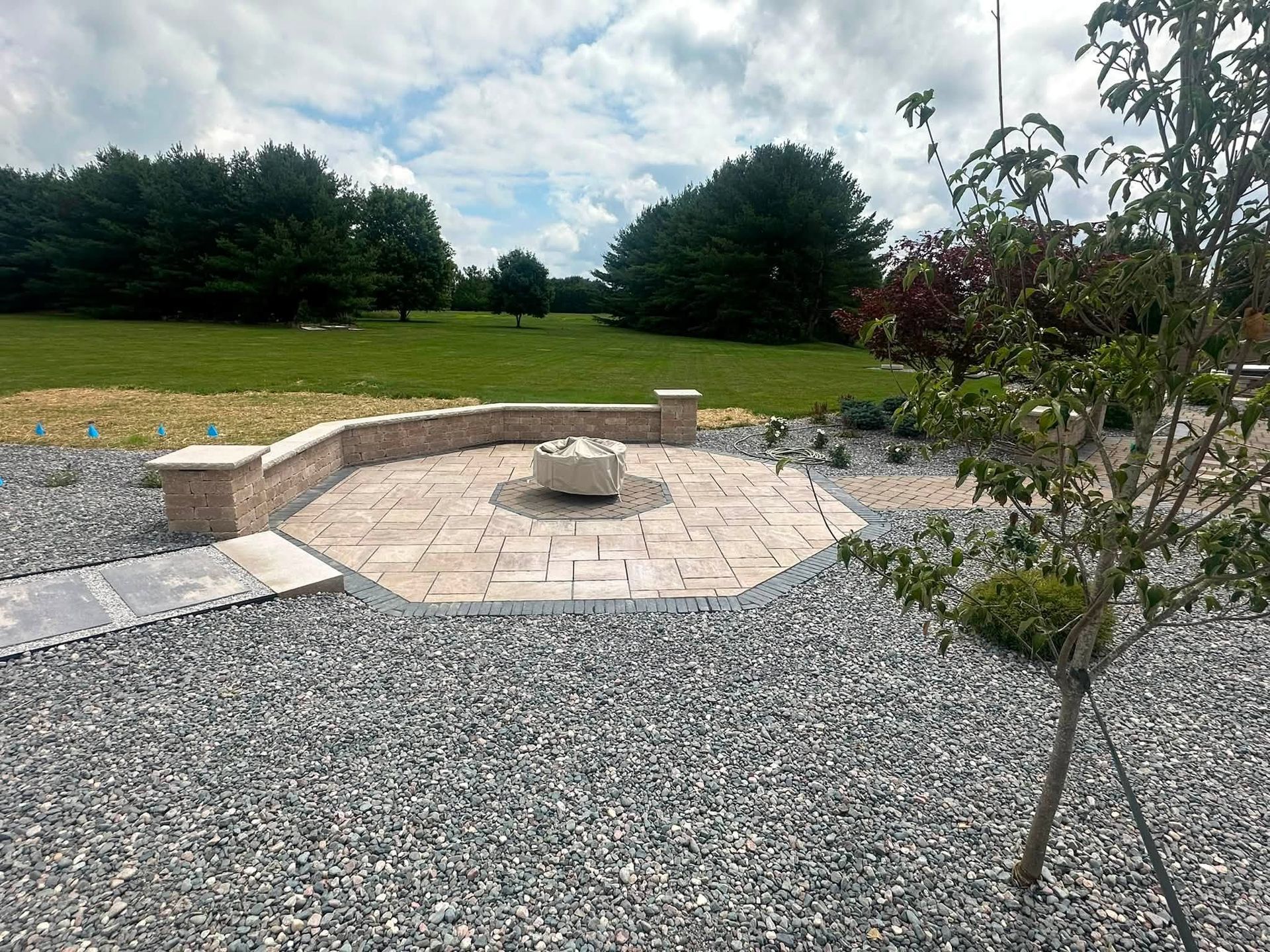 Stone patio with curved bench and fire pit in a grassy backyard, with gravel and a small tree.