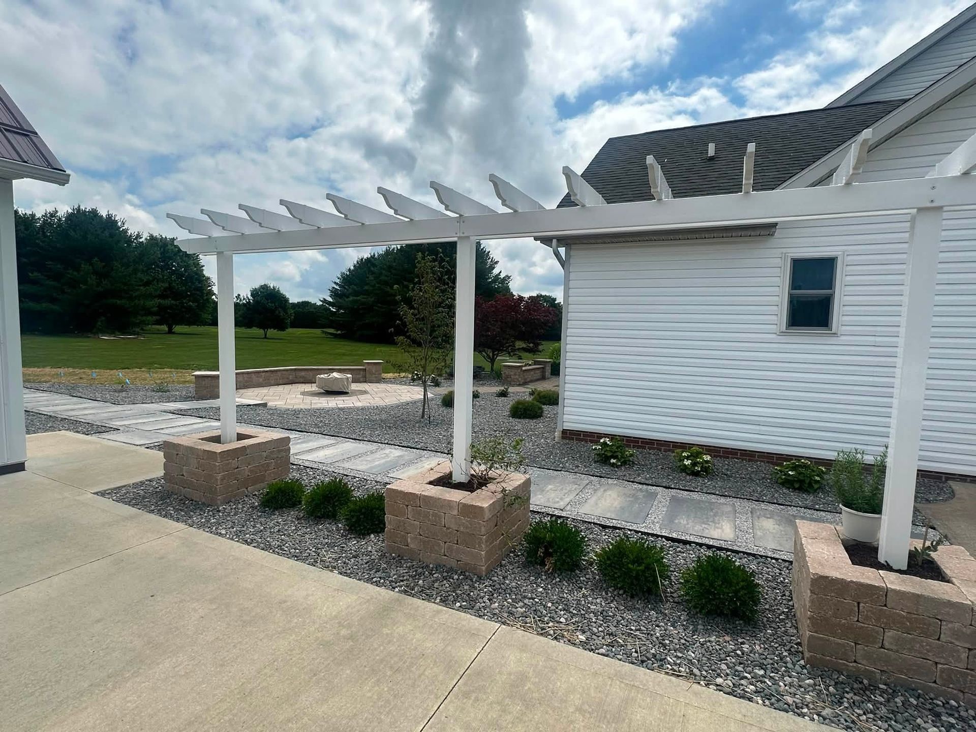 White pergola with brick posts beside a white house and gravel landscaping under a cloudy sky