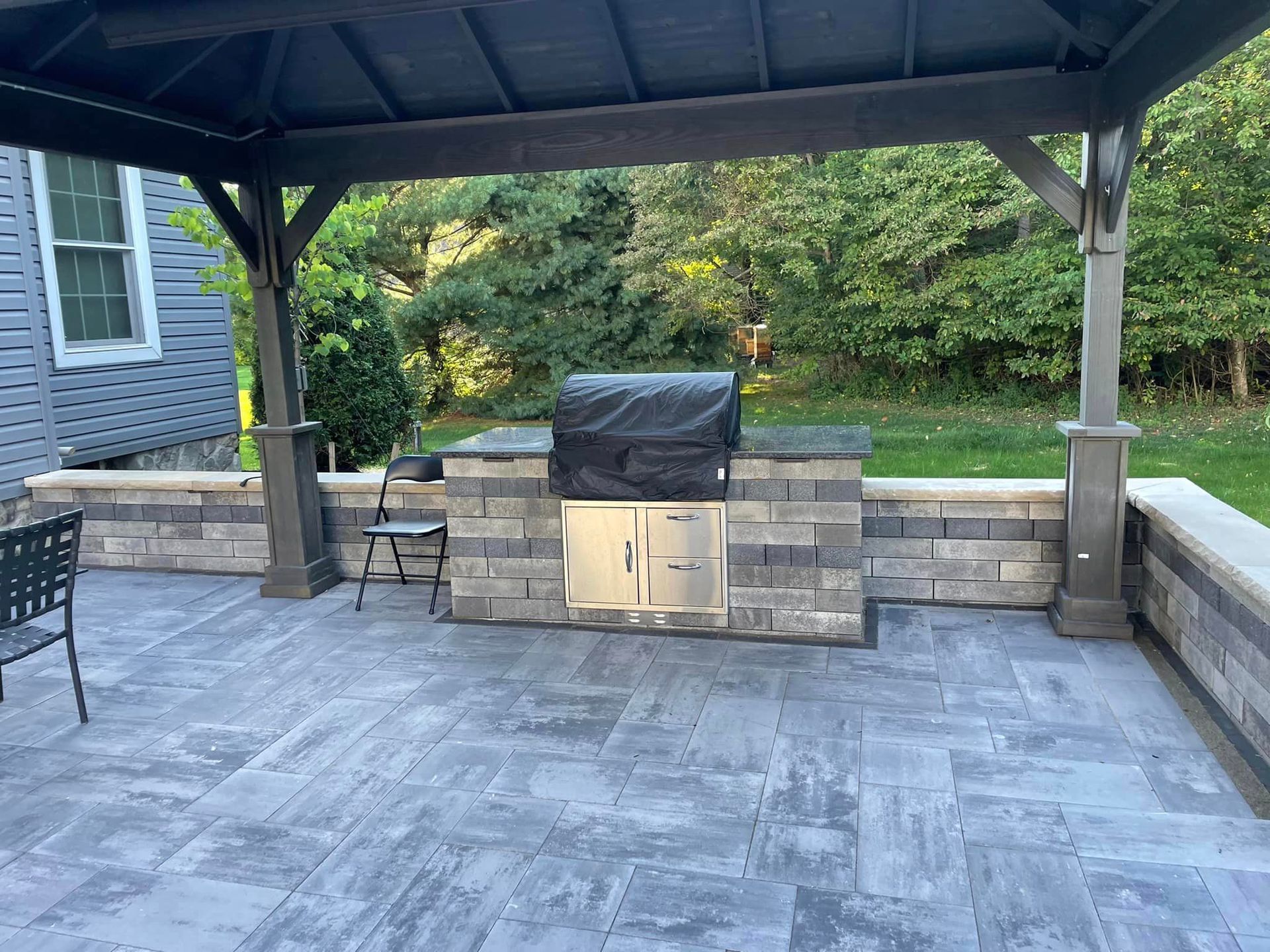 Covered patio with stone grill station and gray paver flooring overlooking a landscaped yard
