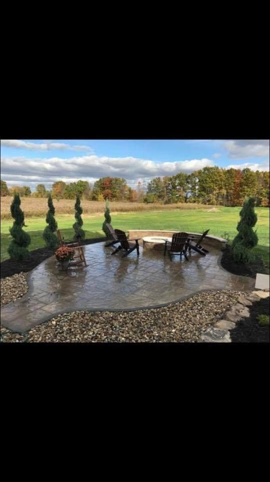 Patio with black chairs and table beside a grassy field, lined with trimmed evergreens under a cloudy sky